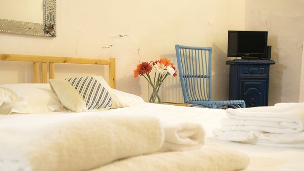 Uma cama bem arrumada com almofadas e toalhas dobradas, uma cadeira, uma mesa pequena com uma televisão e um vaso com flores.