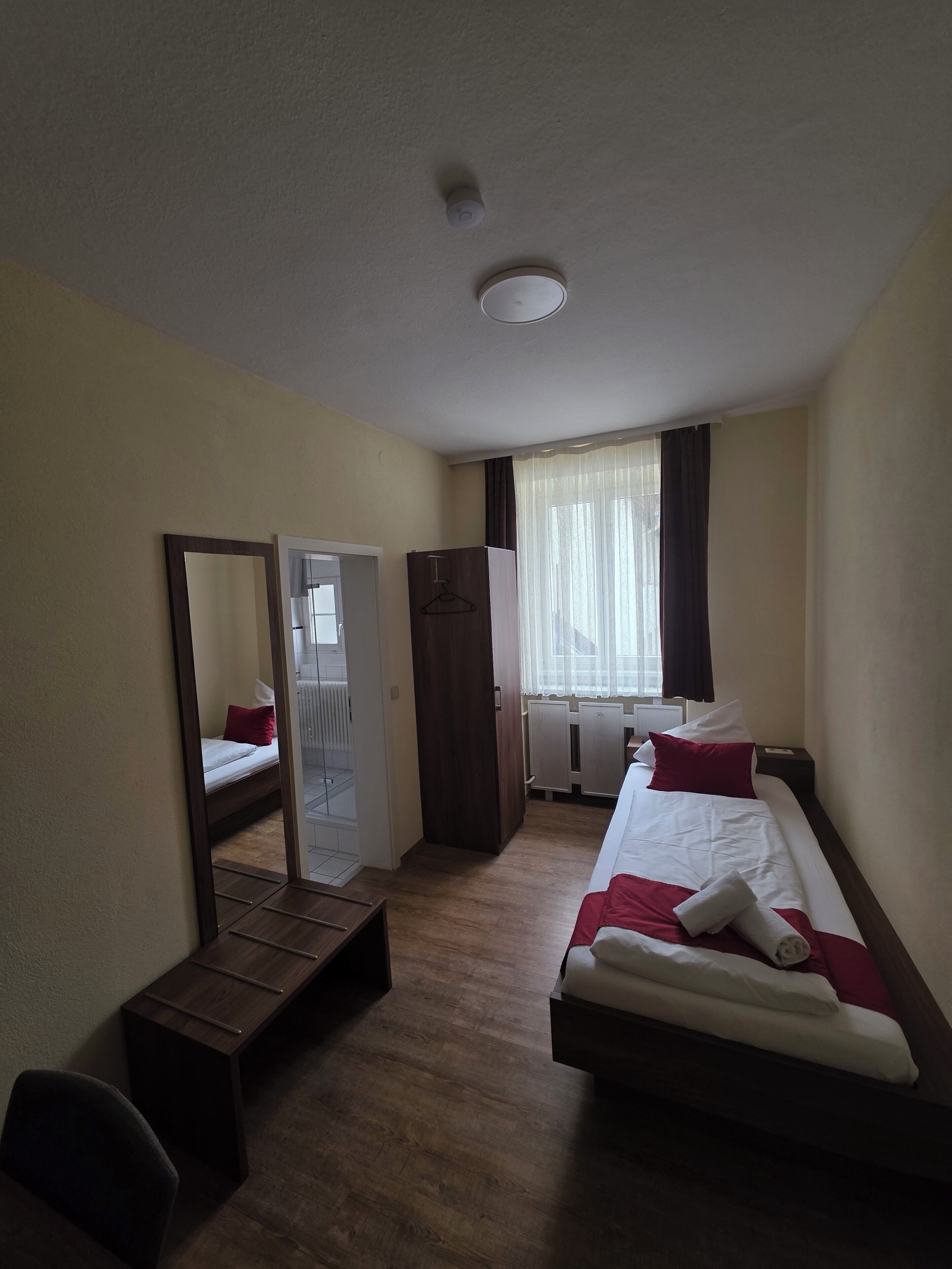 A hotel room with a bed, a wardrobe, a mirror, a desk, and a window with curtains.