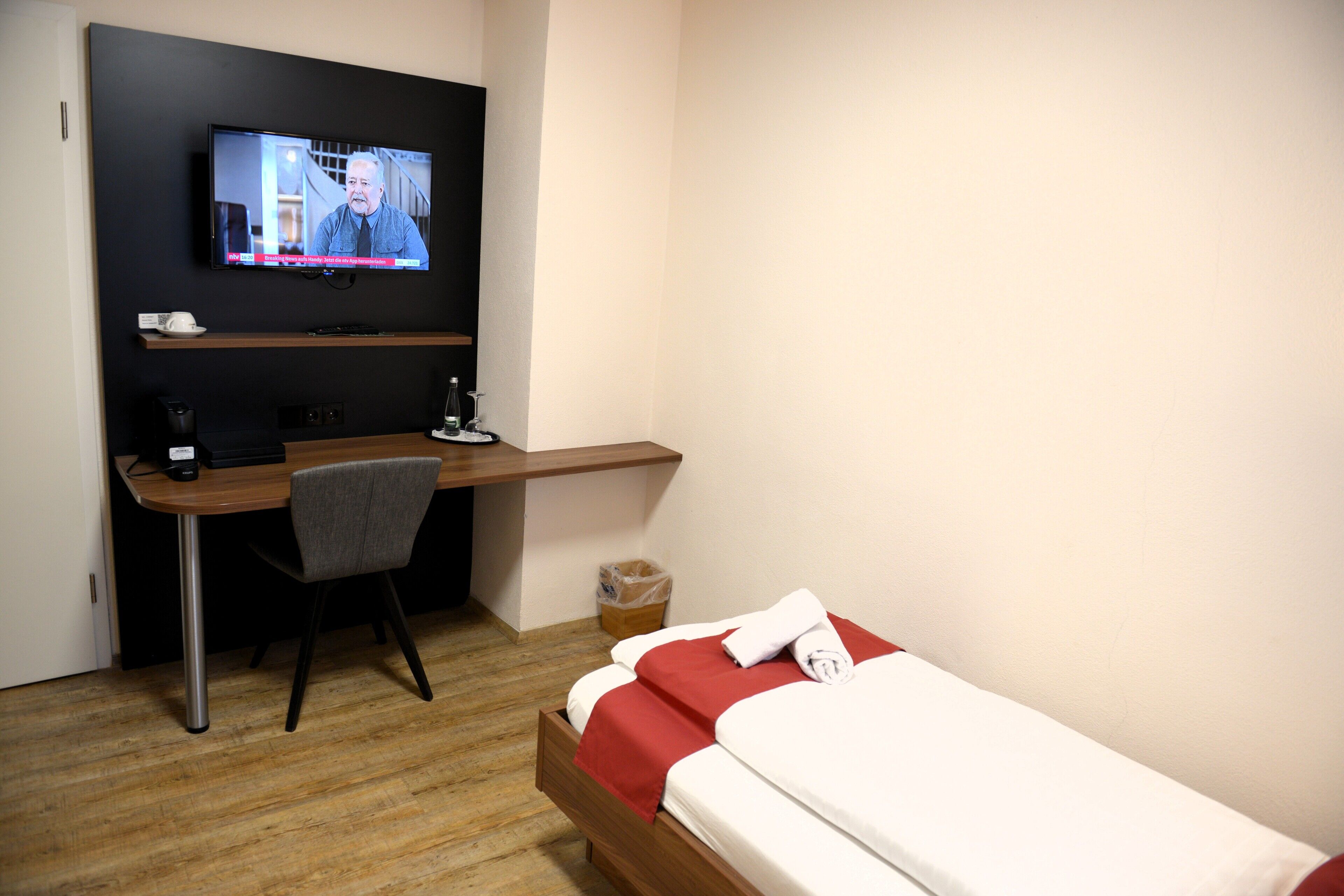A hotel room with a bed, a desk with a chair, a television, and a trash bin.