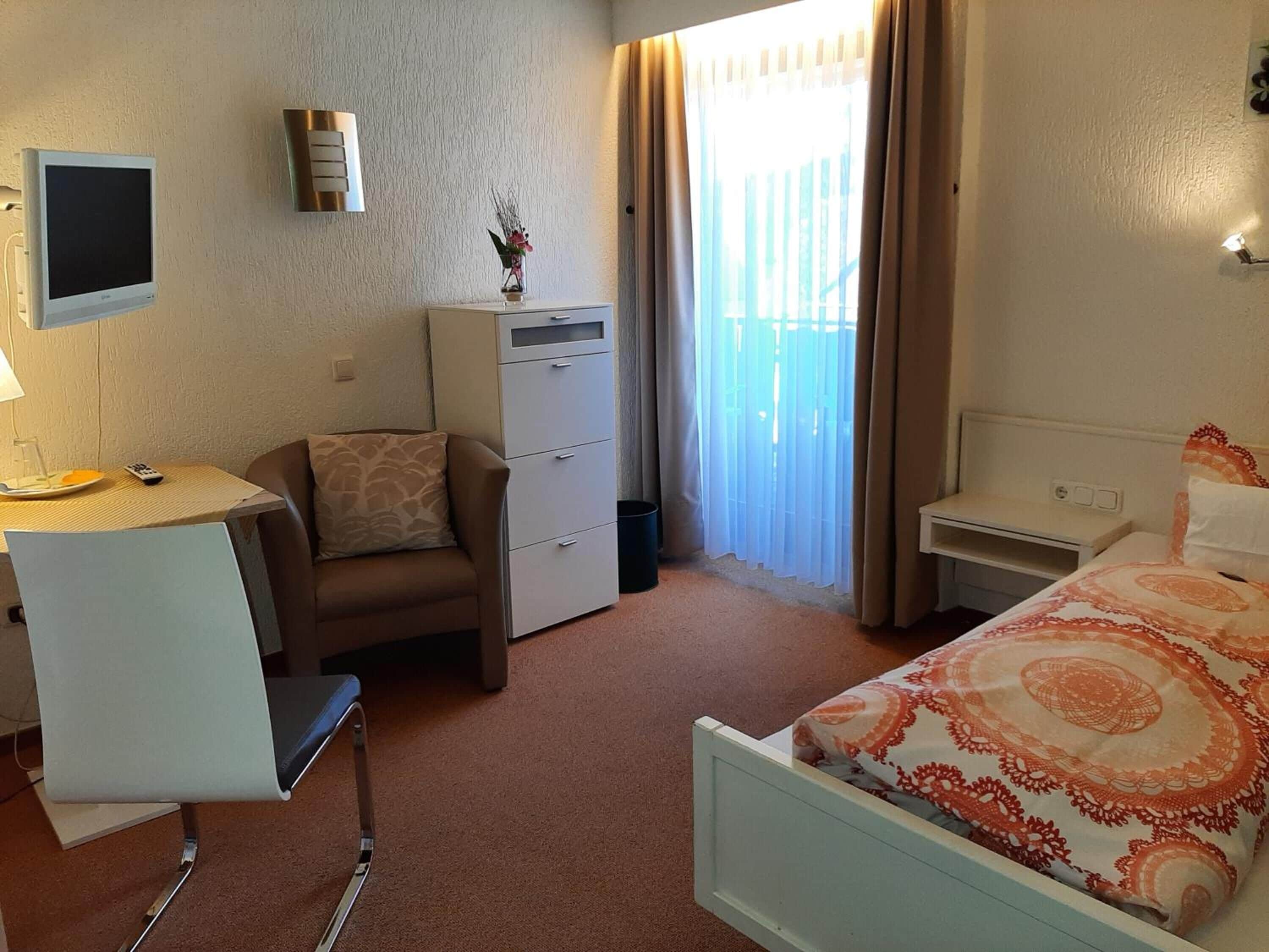 Une chambre d’hôtel comprenant un lit, un bureau avec une chaise, un petit placard et une fenêtre avec des rideaux.