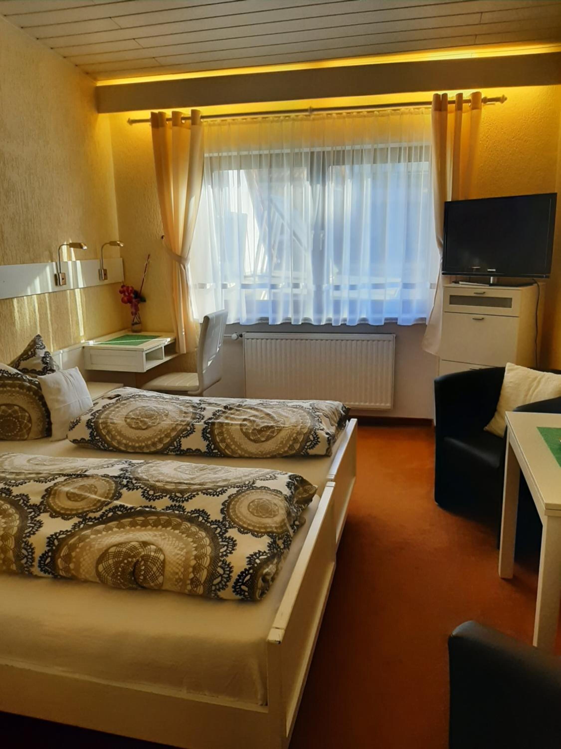 Habitación de hotel con cama, escritorio, silla, televisor y ventana con cortinas.