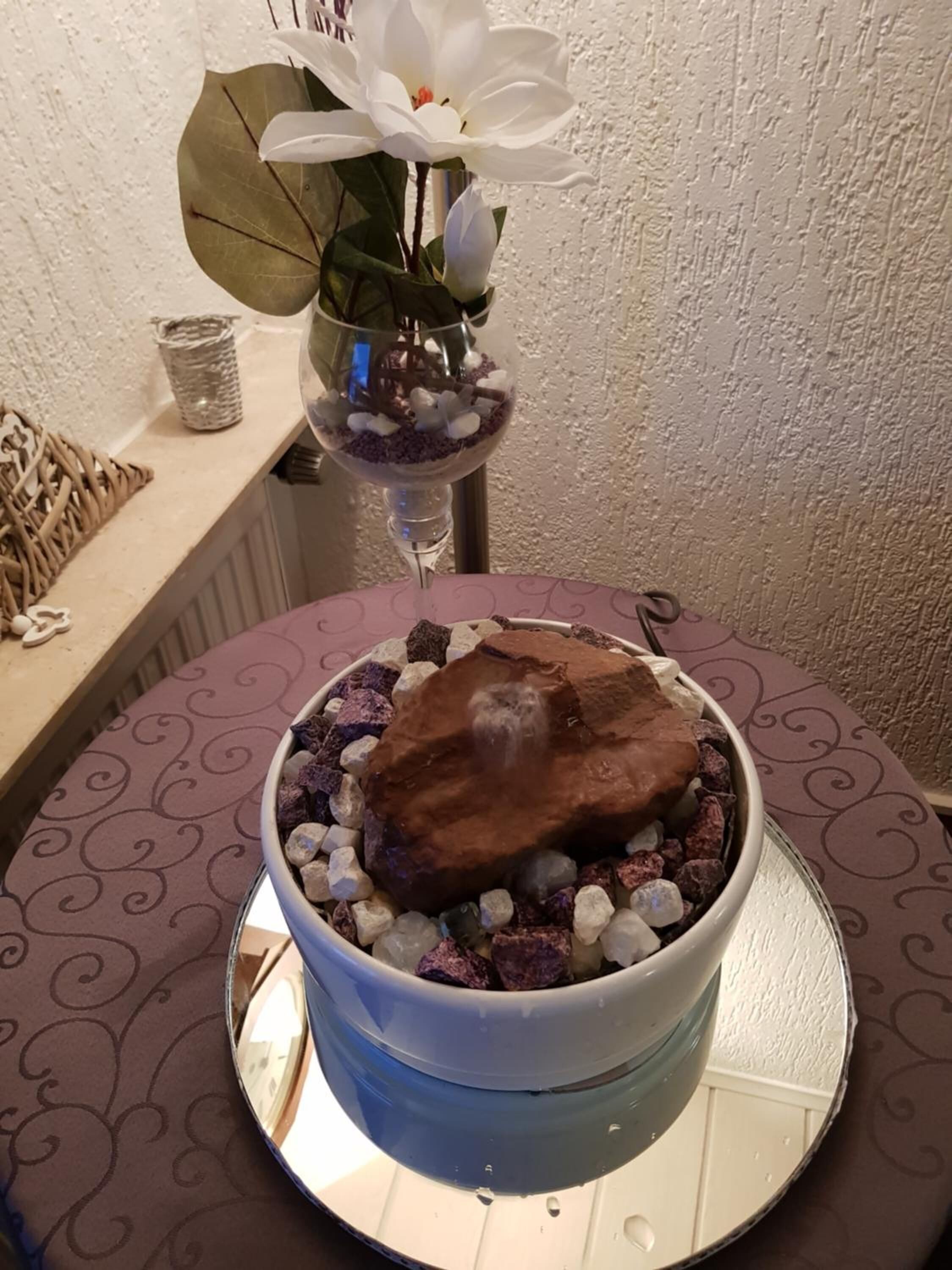 Un cuenco decorativo con un trozo de chocolate encima, rodeado de piedras pequeñas y una flor blanca en un florero de vidrio.