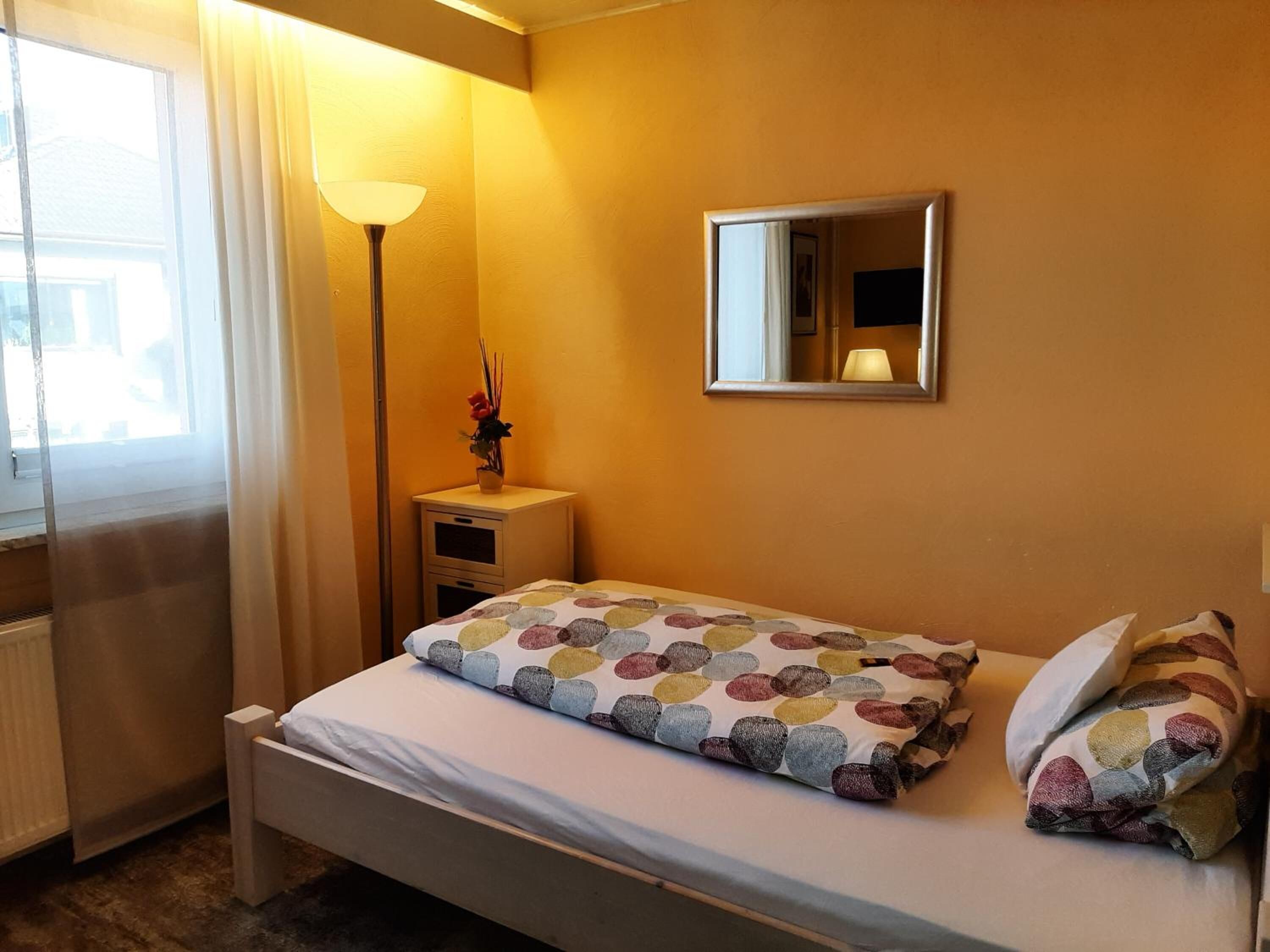 A bedroom with a bed, a bedside table, a lamp, a mirror, and a window with curtains.