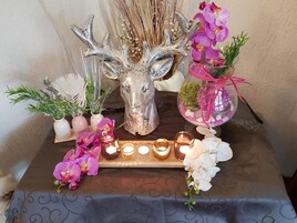 Ein Tisch mit dekorativem Mittelstück, das einen silbernen Hirschschädel zeigt, umgeben von kleinen Topfpflanzen und rosa Orchideen.