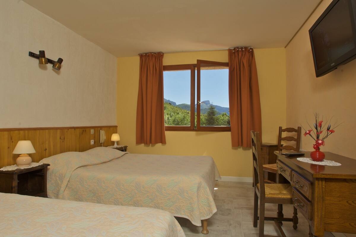 Ein Hotelzimmer mit zwei Betten, einem Holztisch, einem Stuhl, einem Fernseher und einem Fenster mit Vorhängen.