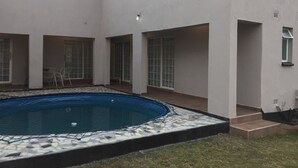 Une piscine dont le bord est revêtu de carrelage, entourée d’une terrasse couverte aménagée avec des chaises et une table.