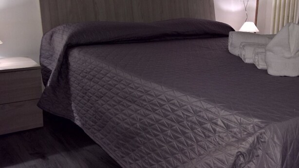 Een netjes opgemaakt bed met een donge deken, een bedlampje en een opgevouwen handdoek op het nachtkastje.