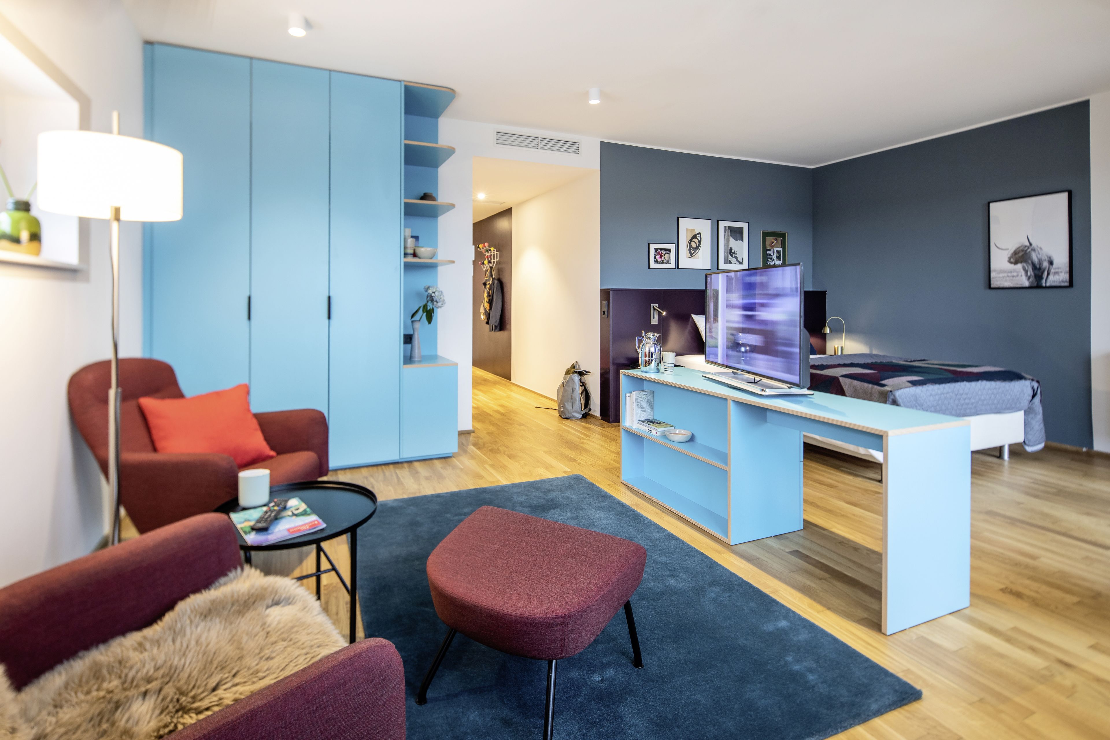 Una sala de estar moderna con un sofá azul, un sillón rojo, una mesita y un reposapiés rojo. Se observa un escritorio con una computadora y una estantería.