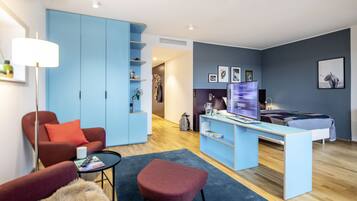Una sala de estar moderna con un sofá azul, un sillón rojo, una mesita y un reposapiés rojo. Se observa un escritorio con una computadora y una estantería.