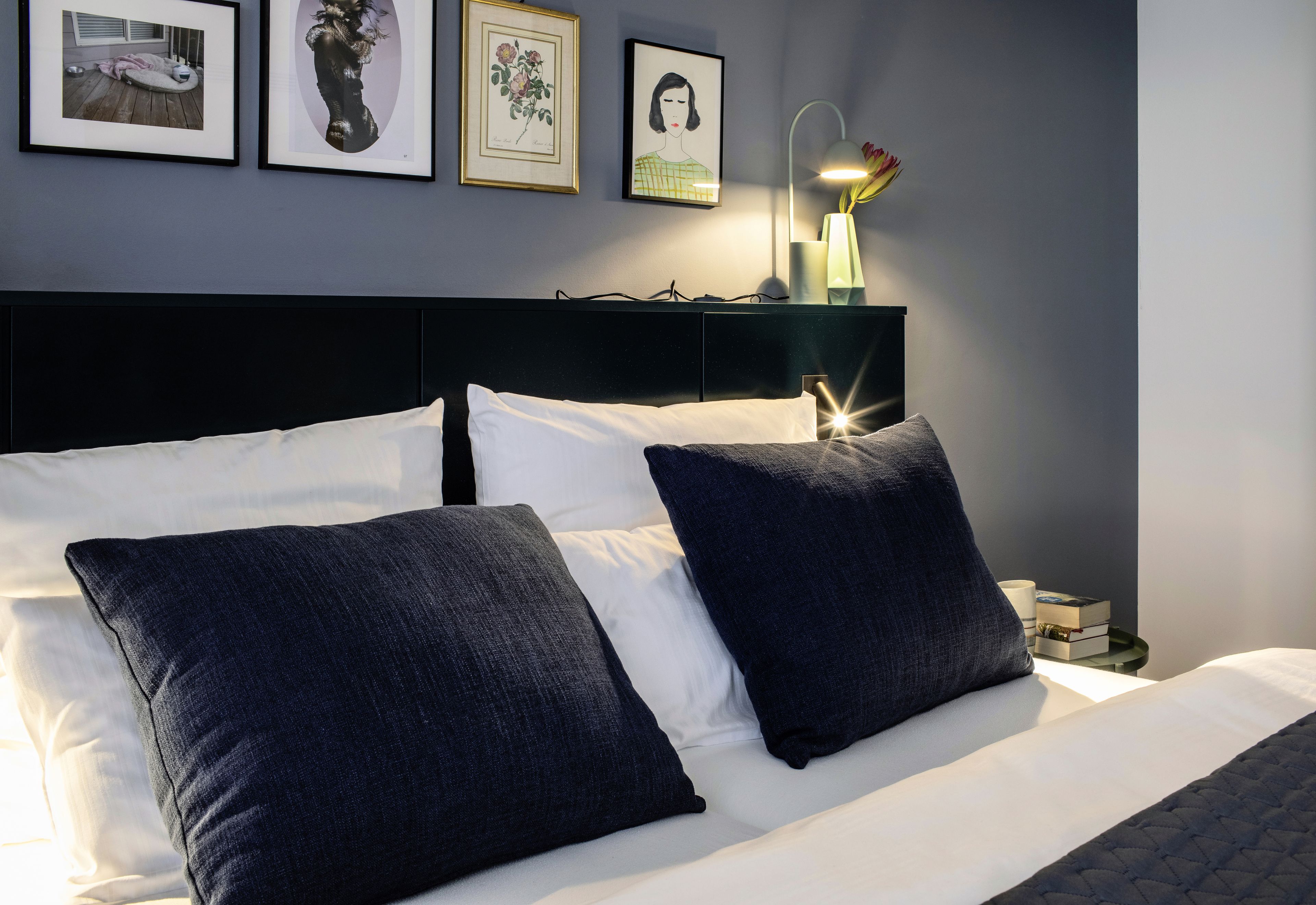 Una cama bien tendida con almohadas blancas y azul oscuro, un cabecero negro y una mesita de noche con una lámpara y un jarrón de flores.
