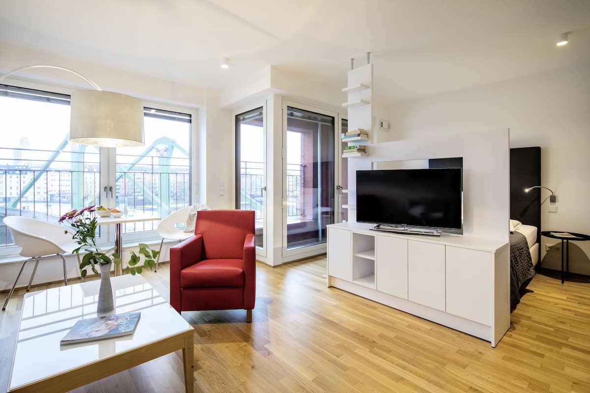 Ein modernes Wohnzimmer mit einem Flachbildfernseher, einem roten Sessel, einem weißen Couchtisch mit einer Vase und Blick auf die Stadt durch große Fenster.
