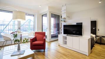 A modern living room with a flat-screen TV, a red armchair, a white coffee table with a vase, and a city view through large windows.