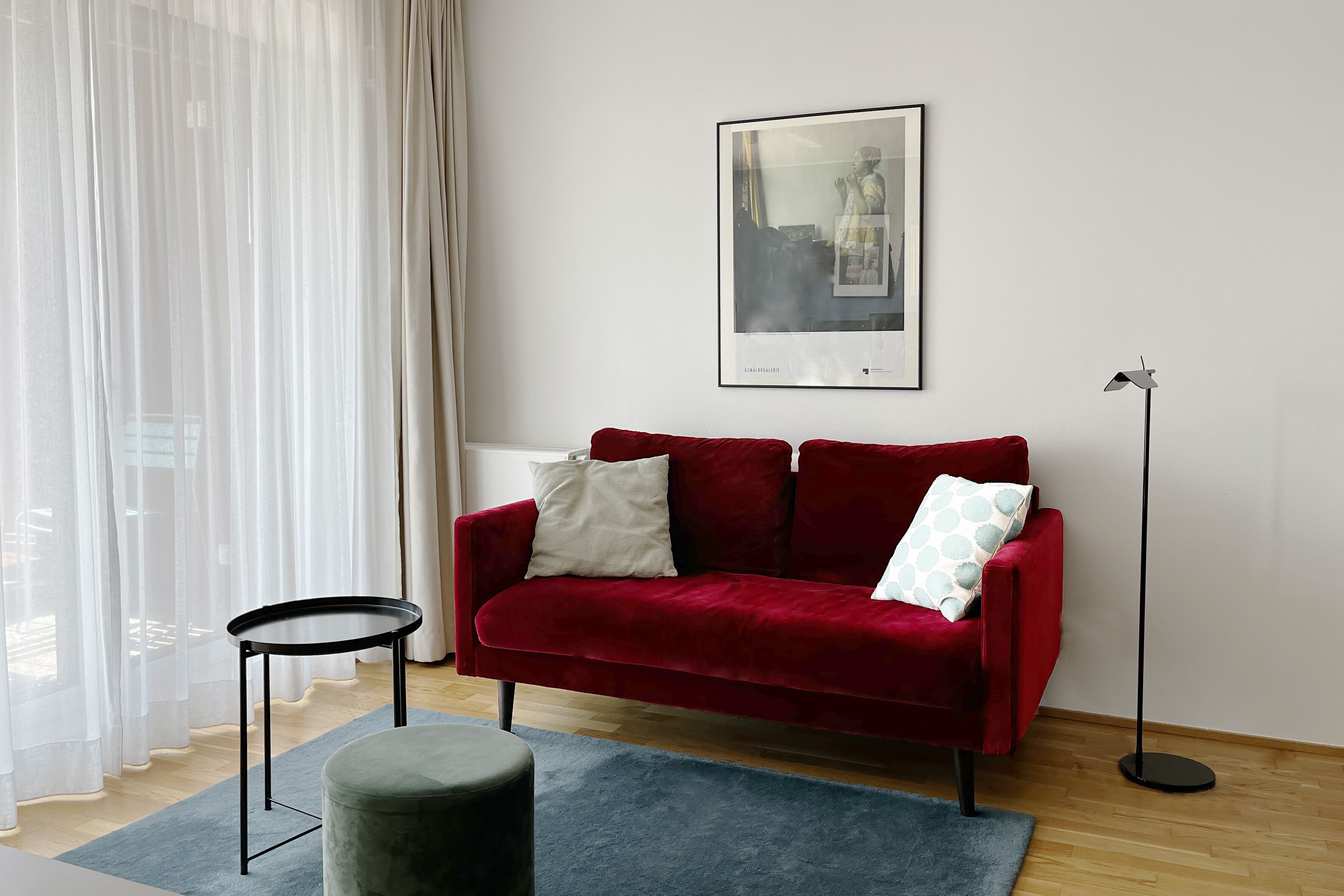 A red velvet sofa with two pillows, a small round table, a blue rug, and a floor lamp.