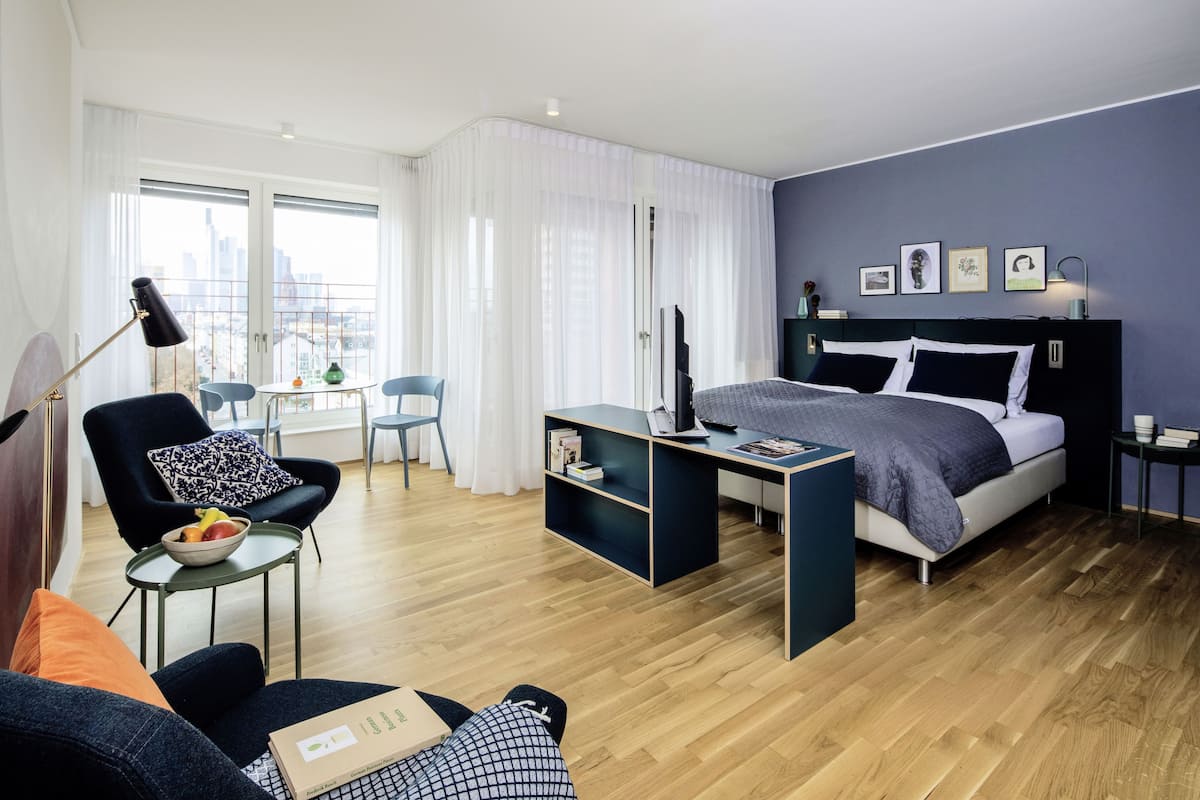 Ein modernes Hotelzimmer mit Bett, Schreibtisch, Stuhl, einem kleinen Tisch mit Obstschale und Blick auf die Stadt durch große Fenster.