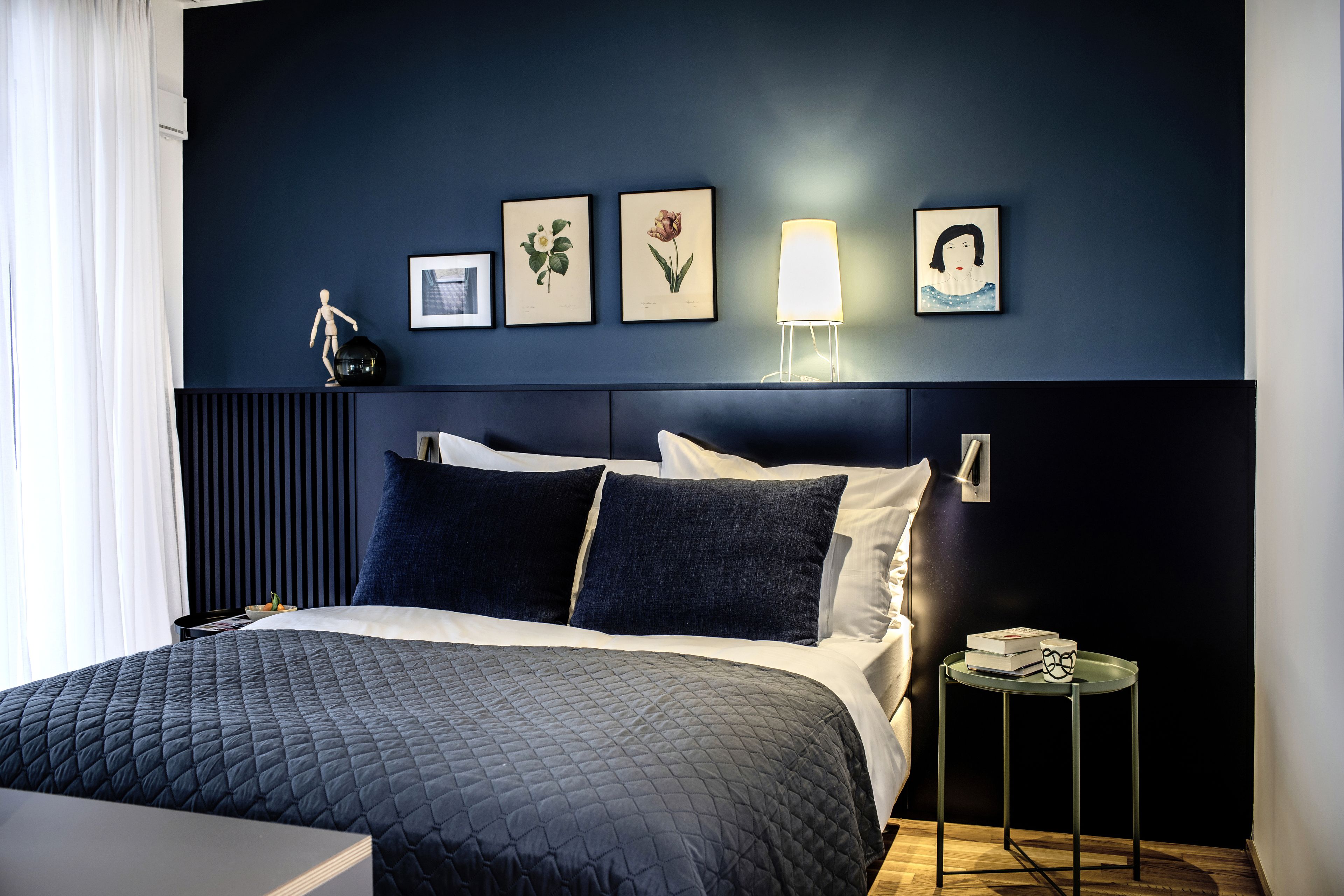 Een moderne hotelkamer met een donkerblauwe accentmuur, een groot bed met een gewatteerd dekbedovertrek, een nachtkastje met een lamp en ingelijste botanische prints.