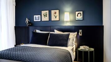 Een moderne hotelkamer met een donkerblauwe accentmuur, een groot bed met een gewatteerd dekbedovertrek, een nachtkastje met een lamp en ingelijste botanische prints.