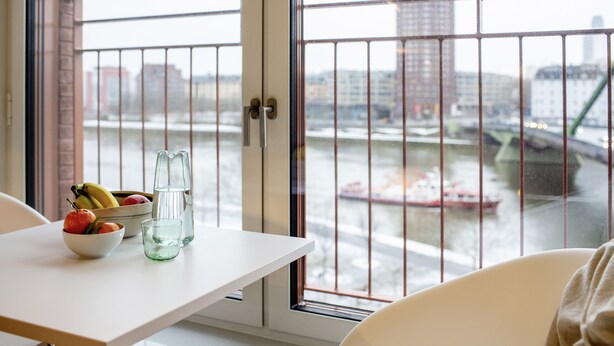 A table with a bowl of fruit, a glass, and a pitcher, with a view of a river and buildings through a window with metal bars. - THE FLAG Oskar M. (Frankfurt)