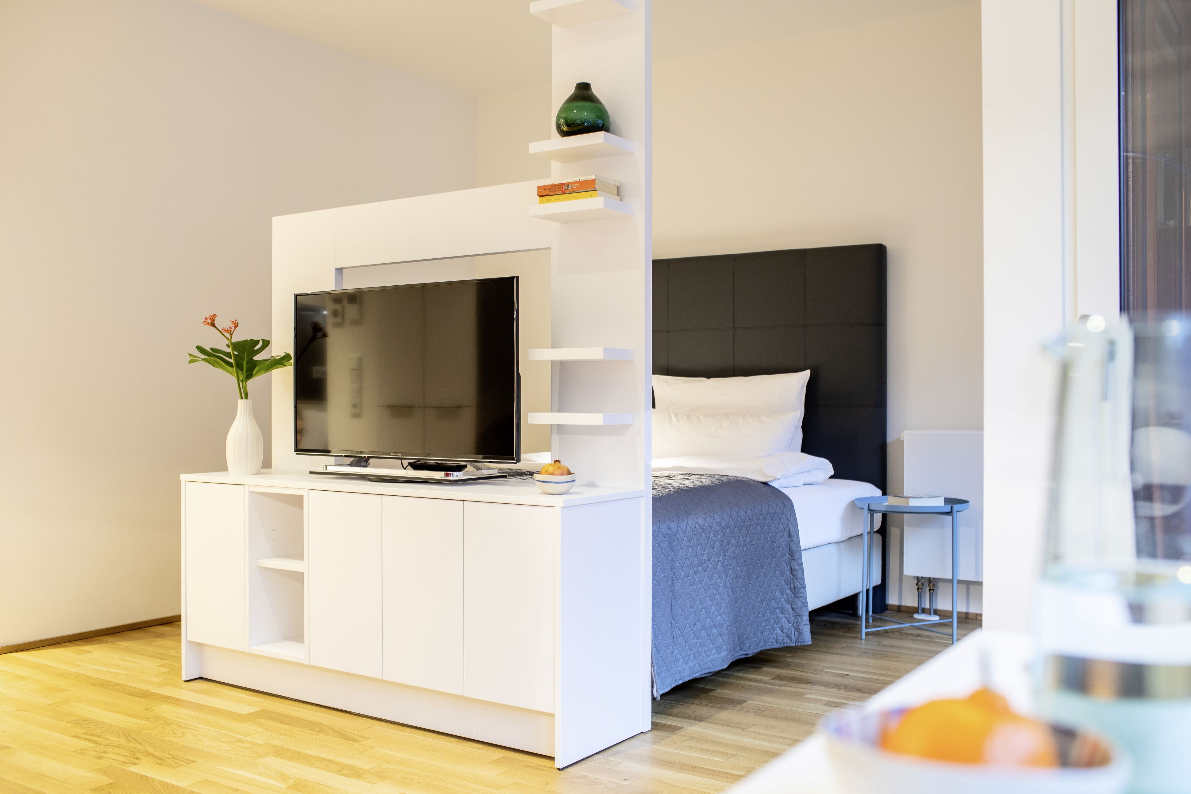A modern bedroom with a bed, a TV on a white console, a small table, and a vase with flowers.