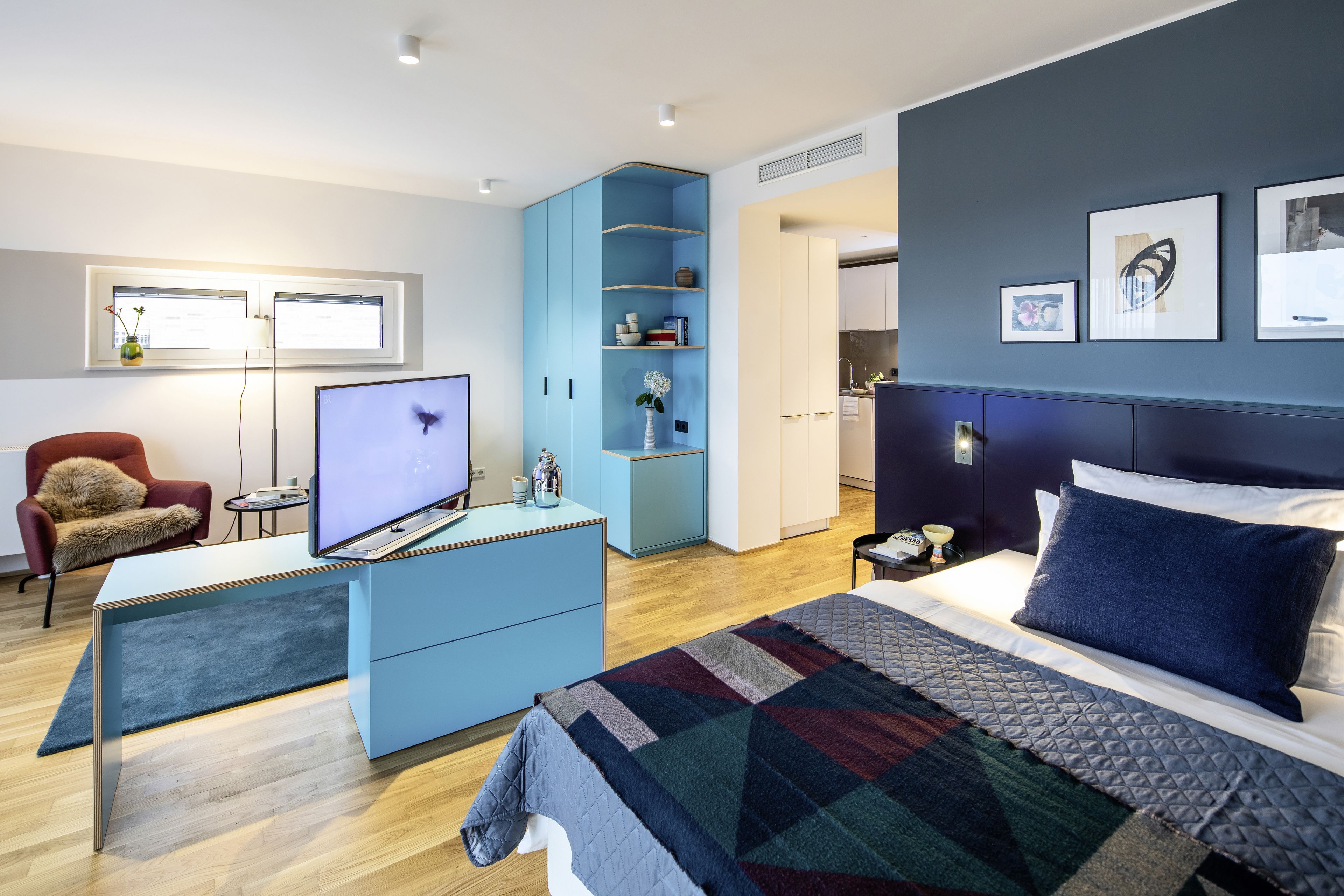 Habitación de hotel moderna con una cama grande, un escritorio con televisor de pantalla plana, un armario azul y una zona de comedor con vistas a la ciudad.