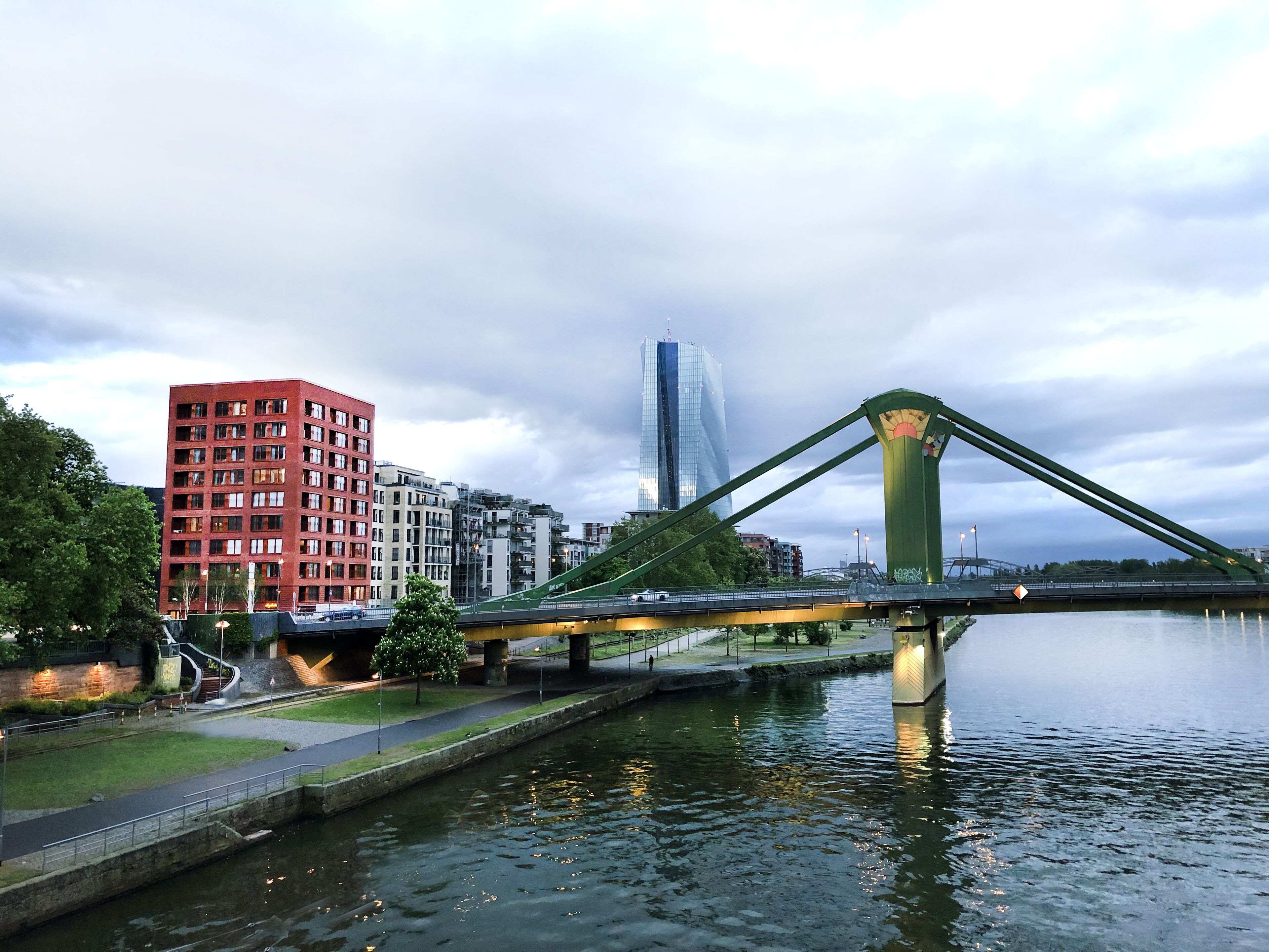 Eine Brücke über einem Fluss, im Hintergrund ein modernes Gebäude und ein rotes Backsteinhaus.