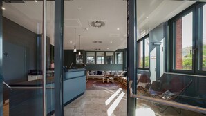 A modern interior with a glass partition, a seating area with sofas and a coffee table, and a kitchen area with a counter and appliances. - THE FLAG Oskar M. (Frankfurt)