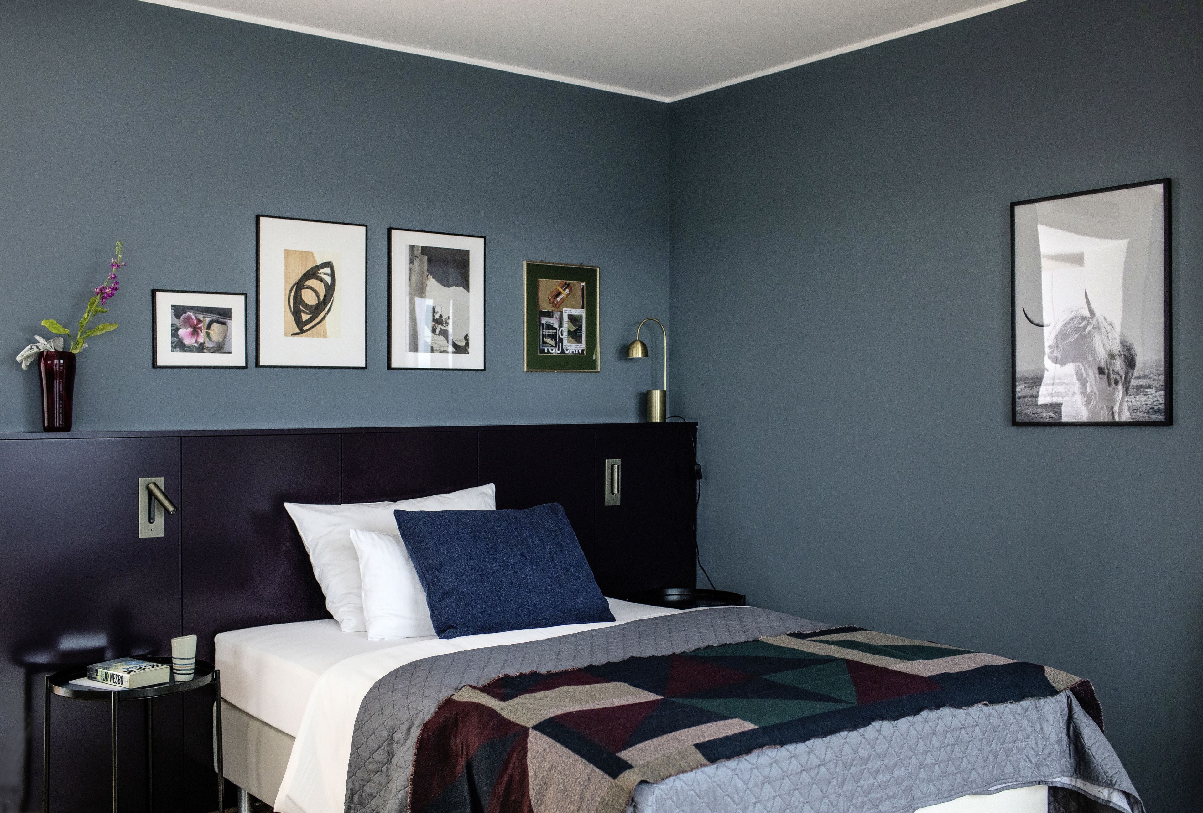 Ein Schlafzimmer mit einem Bett, einem Nachttisch, einer Wandlampe und gerahmten Kunstwerken.
