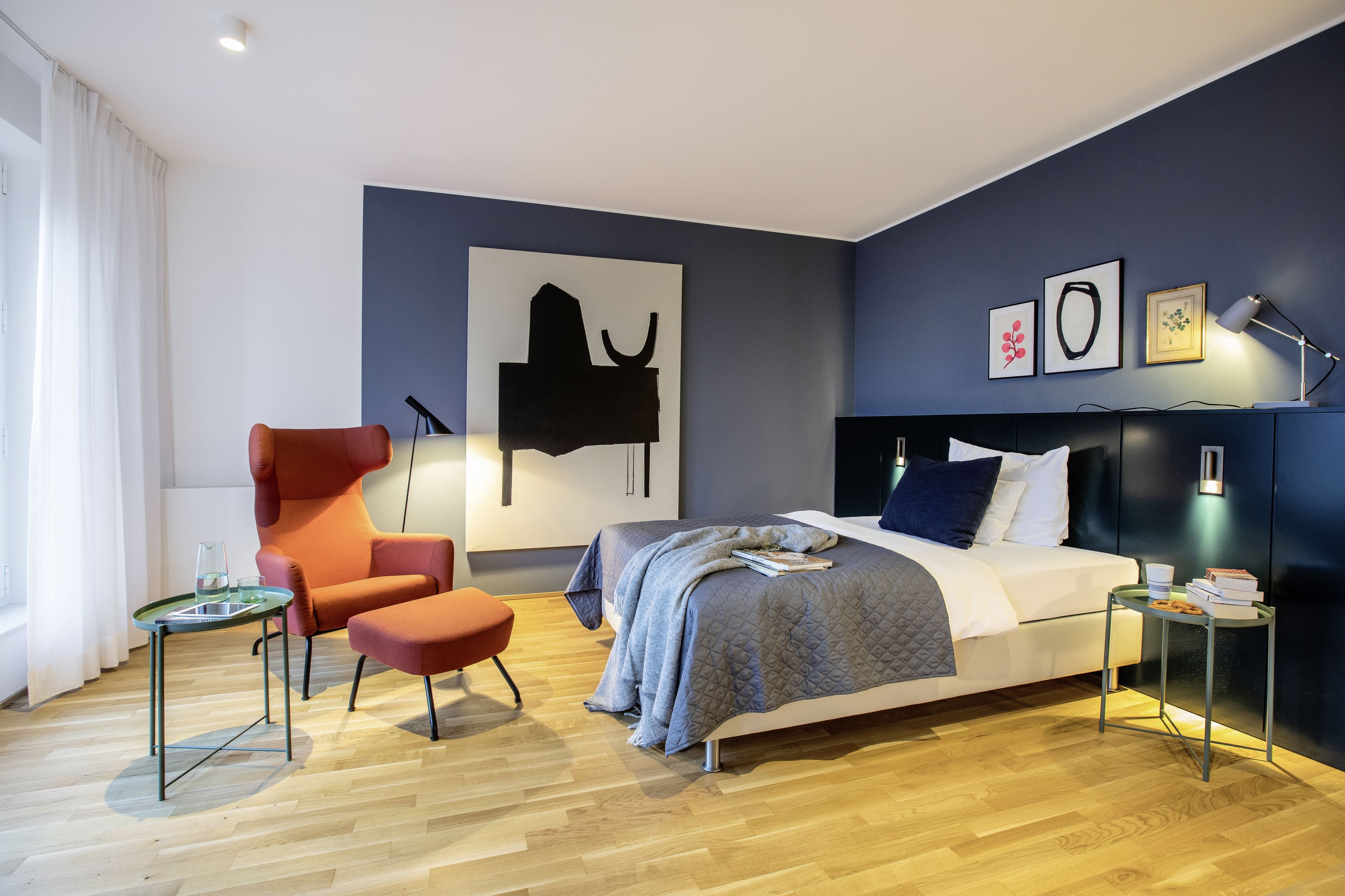 A modern bedroom with a bed, a chair, a small table, and artwork on the wall.