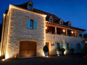 A stone building with a wooden door, potted plants, and a balcony with lights. - Hôtel Bon Encontre (Sarlat-la-Canéda)