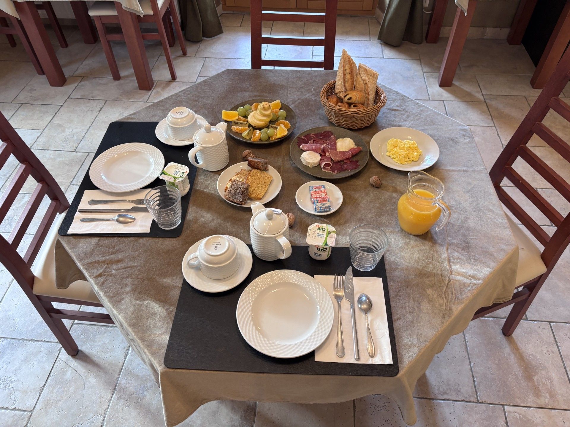 A breakfast setting with a table set for four, including plates, cutlery, a jug of orange juice, and various breakfast items.