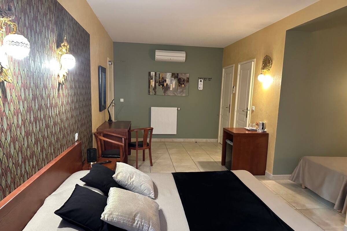 A hotel room with a bed, a desk, a chair, and a small table. There is a window and an air conditioning unit on the wall.