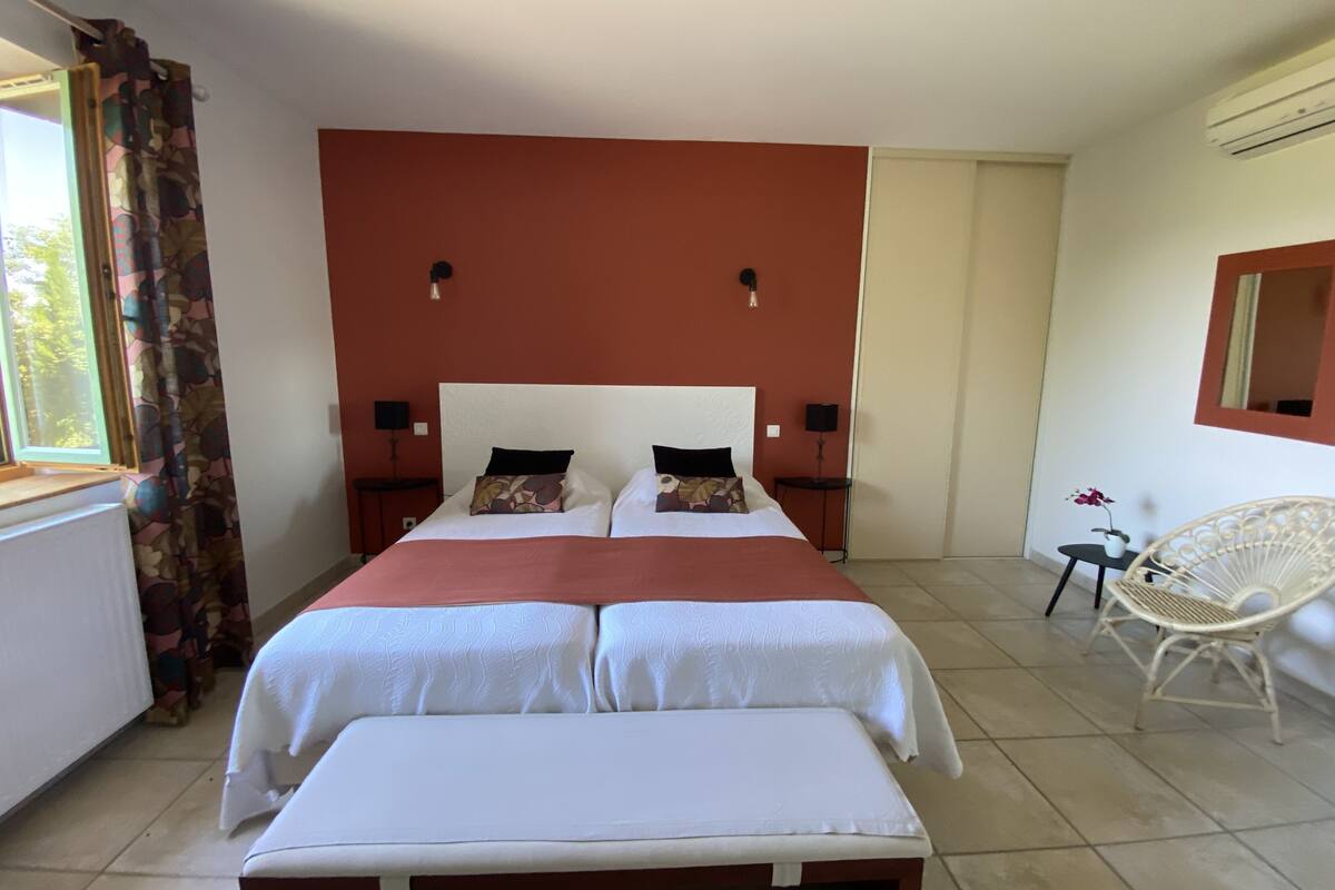 A double bed with a white and red bedspread, two bedside tables with lamps, a white chair, and a window with curtains.