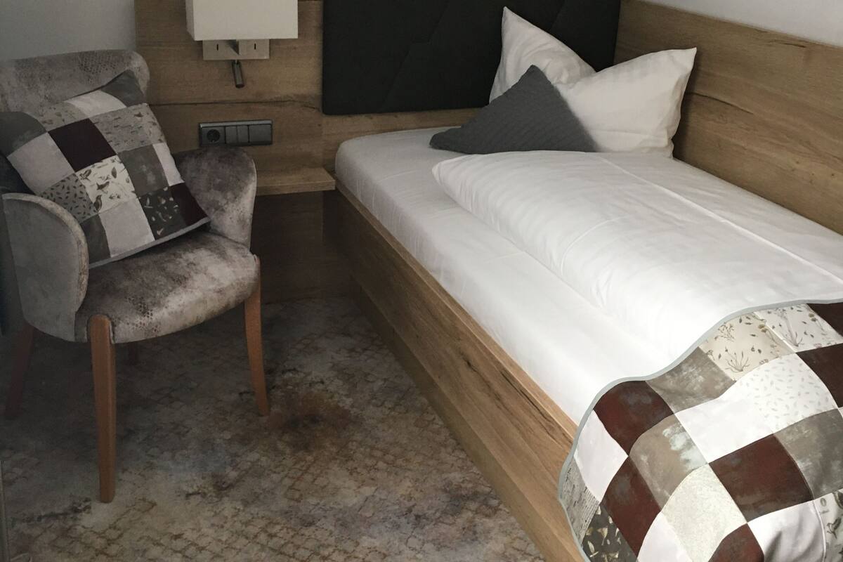 A single bed with a wooden headboard, a bedside table with a lamp, and a chair with a patchwork cushion.