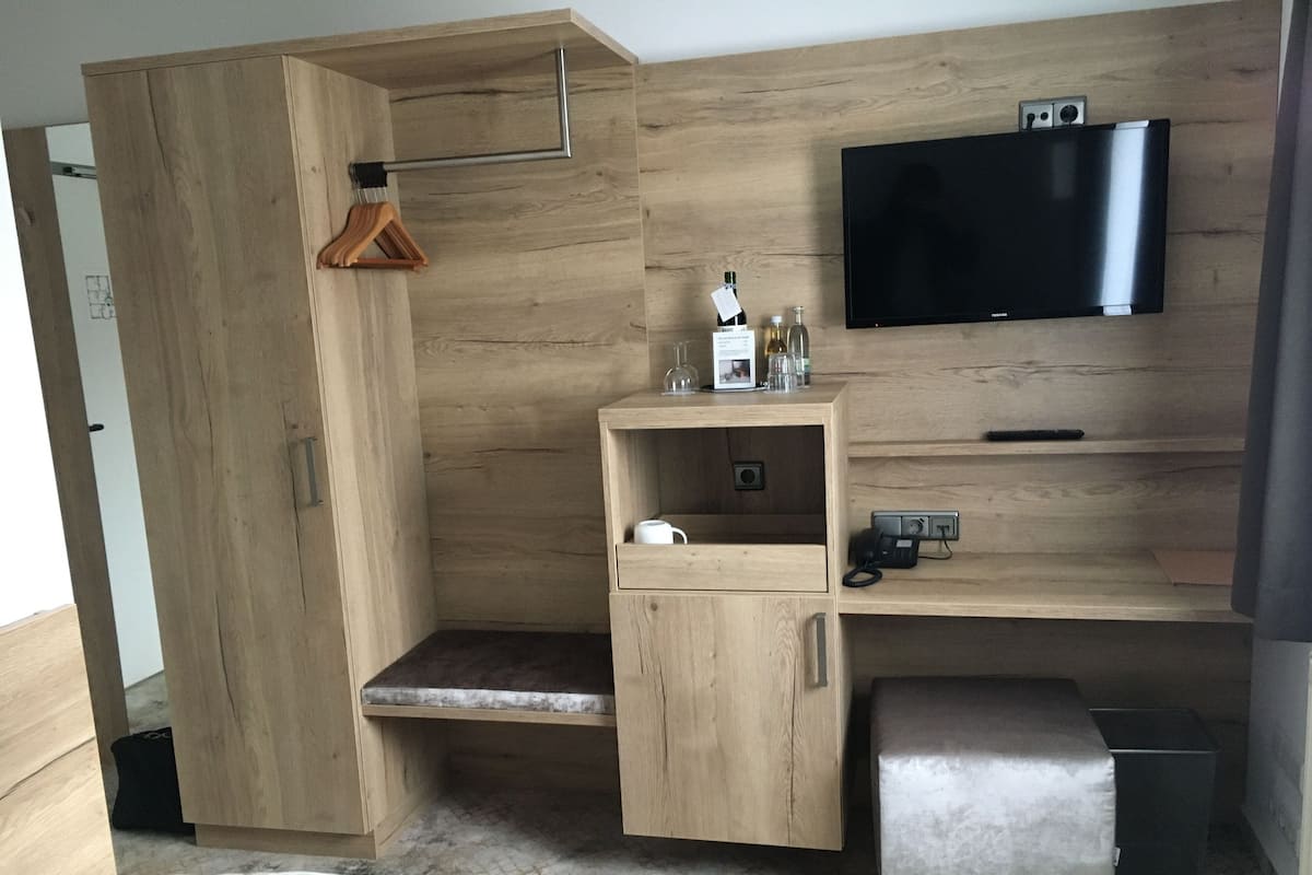 A modern room with a wooden wardrobe, a television mounted on the wall, a small desk with a chair, and a bench.