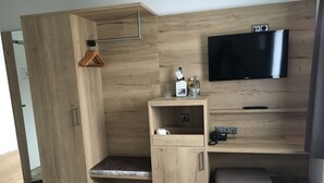 Una habitación moderna con un armario de madera, un escritorio con televisor de pantalla plana, un banco y una mesita con una botella y vasos.