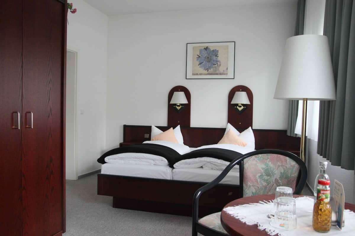 A hotel room with a double bed, a wooden wardrobe, a framed picture on the wall, a table with a lamp, and a bottle of water.