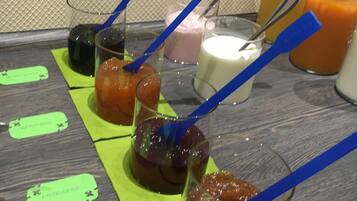 Une table sur laquelle sont disposés divers aliments et boissons, chacun accompagné d’une cuillère bleue et portant une étiquette avec des noms tels que « Aprikoze » et « Bratapfel ».