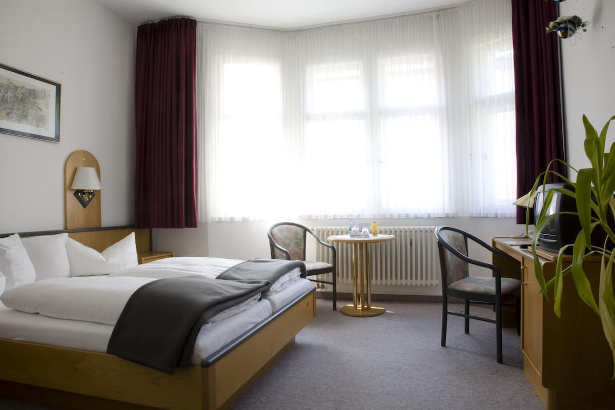 A hotel room with a bed, a desk, a chair, a radiator, and a window with curtains.