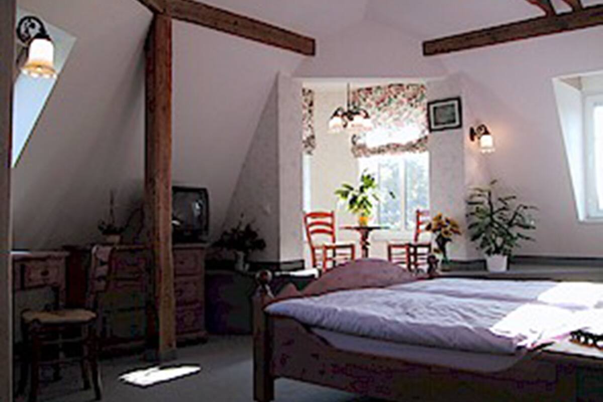 A bedroom with a sloped ceiling, a bed, a desk, and a chair. There are two potted plants and a window with curtains.