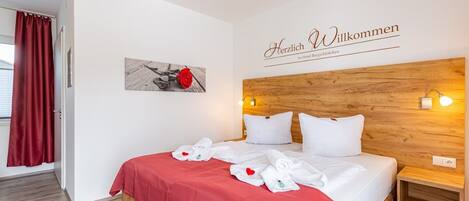 A hotel room with a wooden headboard, white bedding, a red accent on the bed, a welcome sign, and a framed picture of a rose.
