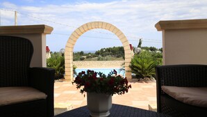 Eine Terrasse mit zwei Korbstühlen, einem Blumentopf mit roten Blüten und Blick auf einen Pool durch einen Bogen.