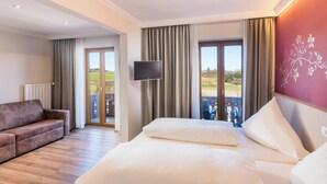 A hotel room with a large bed, a sofa, a TV, and a balcony with a view of the countryside. - Hotel Gasthof Huber (Ebersberg)