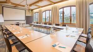 A conference room with long tables, chairs, a projector screen, and large windows with curtains. - Hotel Gasthof Huber (Ebersberg)