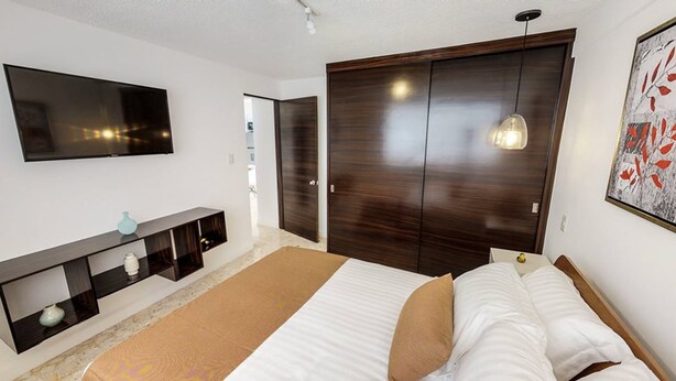 A bedroom with a bed, a TV mounted on the wall, a wardrobe, and a bedside lamp. - Roma Suites (Mexico City)
