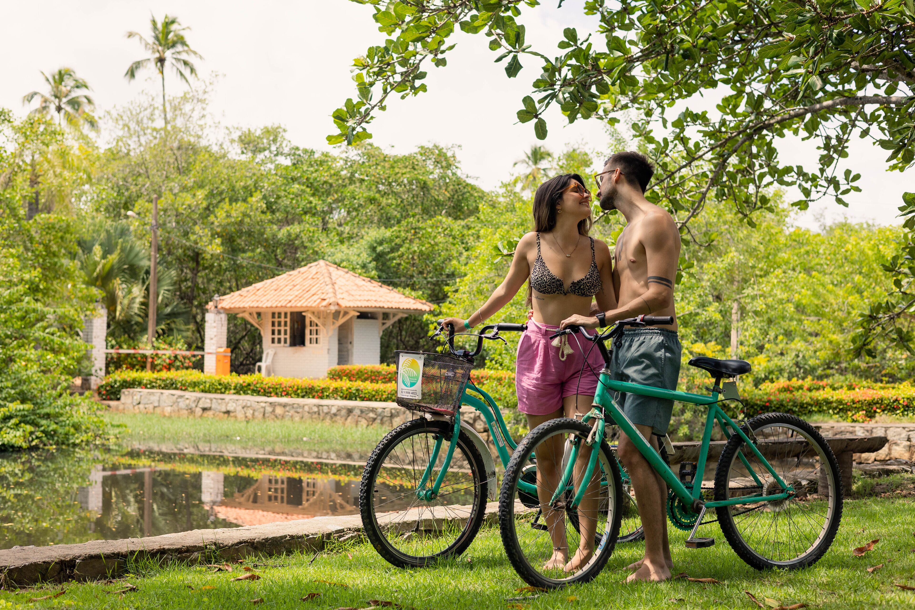 A couple standing by a pond with bicycles, surrounded by lush greenery and a small building in the background.