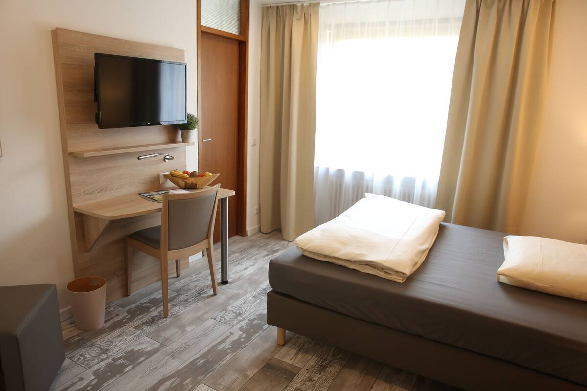 Una habitación de hotel con cama, escritorio con silla, televisor montado en la pared y ventana con cortinas.