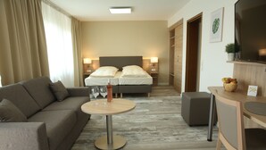A modern hotel room with a sofa, a round table with a bottle and glasses, a bed with two pillows, and a TV on a wooden shelf. - Kemnater Hof Apartments   (Ostfildern)