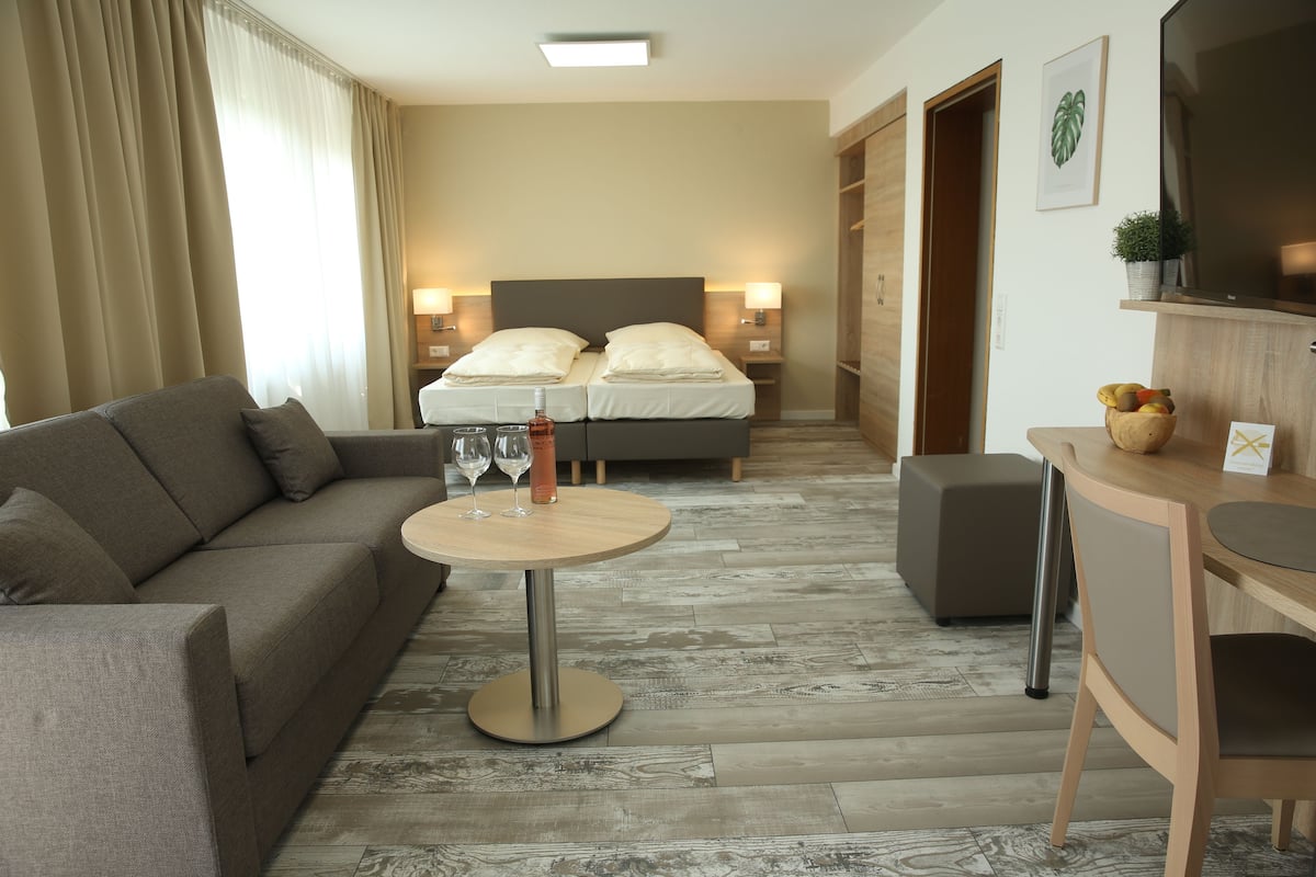 Una habitación de hotel moderna con un sofá, una mesa redonda con una botella y vasos, una cama con dos almohadas y un televisor sobre una estantería de madera.