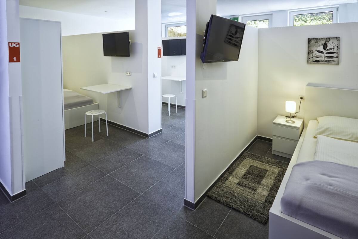 Habitación de hotel con cama, mesita de noche con lámpara, taburete pequeño, escritorio con silla y televisor en la pared.