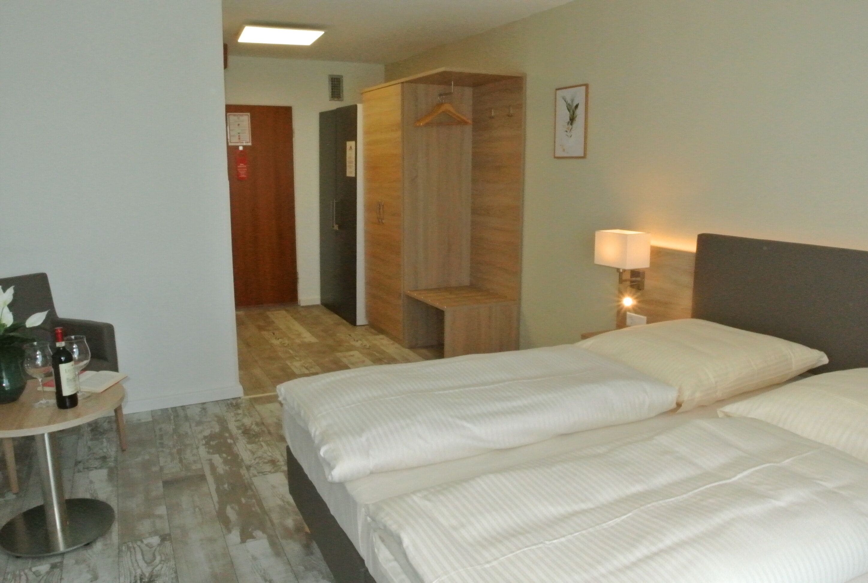 A hotel room with two beds, a wooden wardrobe, a bedside table with a lamp, and a small table with a vase and wine bottle.