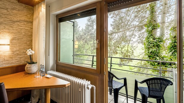 A room with a wooden table, a radiator, and a window with a view of trees and a balcony with chairs. - Kemnater Hof Apartments   (Ostfildern)
